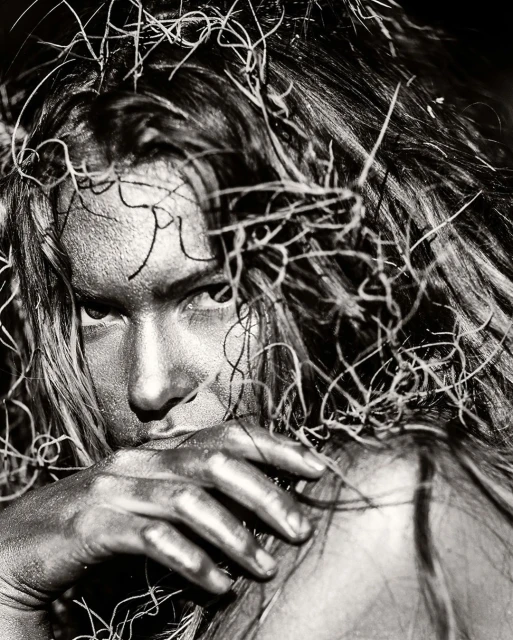 Monochrome close-up of an adult woman with long hair and silver body paint, partially concealing her face with a silver-painted hand.