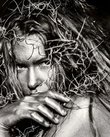 Monochrome close-up of an adult woman with long hair and silver body paint, partially concealing her face with a silver-painted hand.