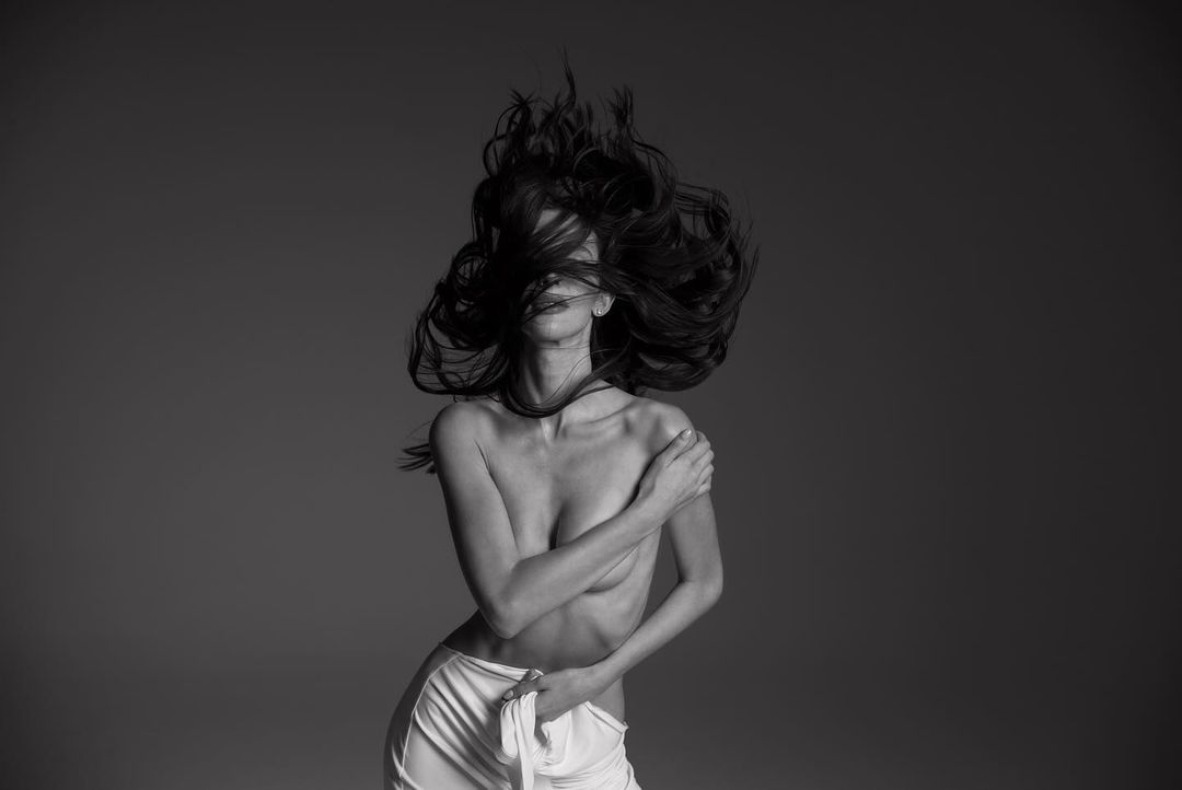 Monochrome portrait of a fair-skinned adult female with long, dark hair, styled in a dynamic pose, wearing a white fabric wrap.