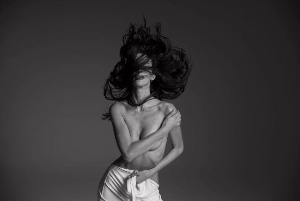 Monochrome portrait of a fair-skinned adult female with long, dark hair, styled in a dynamic pose, wearing a white fabric wrap.