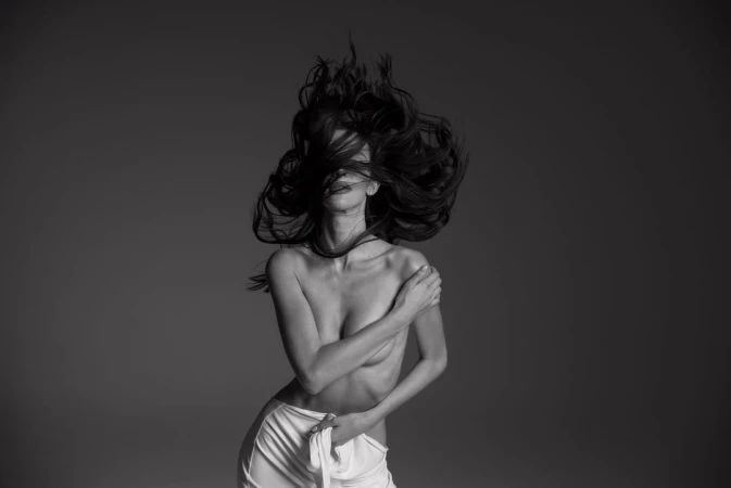 Monochrome portrait of a fair-skinned adult female with long, dark hair, styled in a dynamic pose, wearing a white fabric wrap.