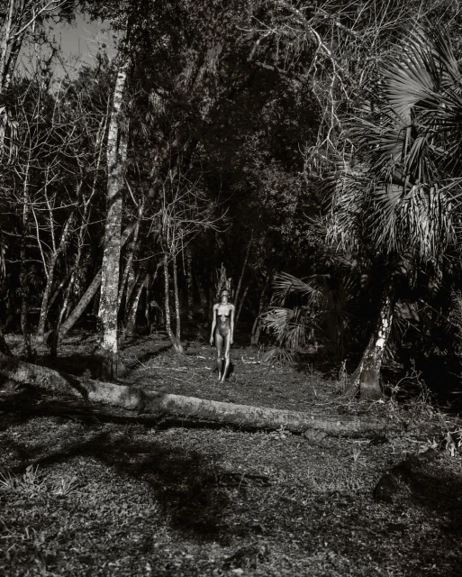 Monochromatic portrait of an adult female in a one-piece bathing suit walking through a densely wooded area with tall trees and thick ground cover.
