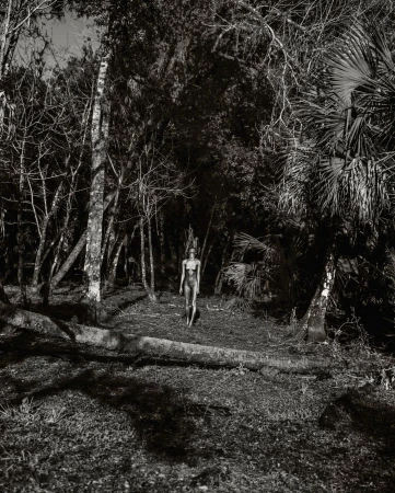 Monochromatic portrait of an adult female in a one-piece bathing suit walking through a densely wooded area with tall trees and thick ground cover.