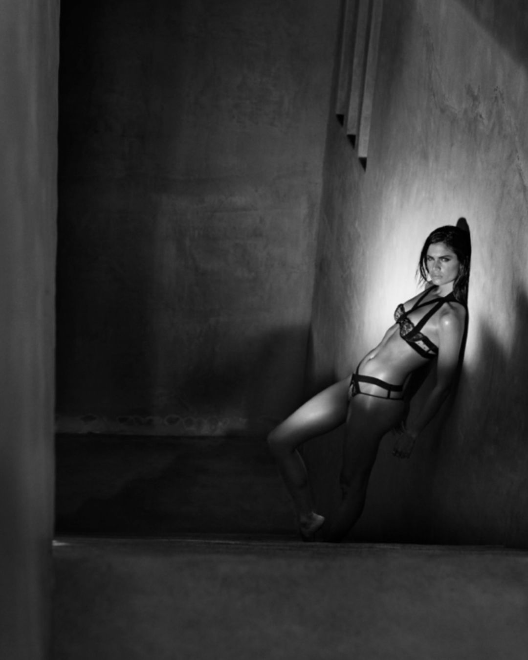 Adult female model in edgy black lingerie leans against a textured wall in a photo studio, emphasizing dramatic light and shadows.
