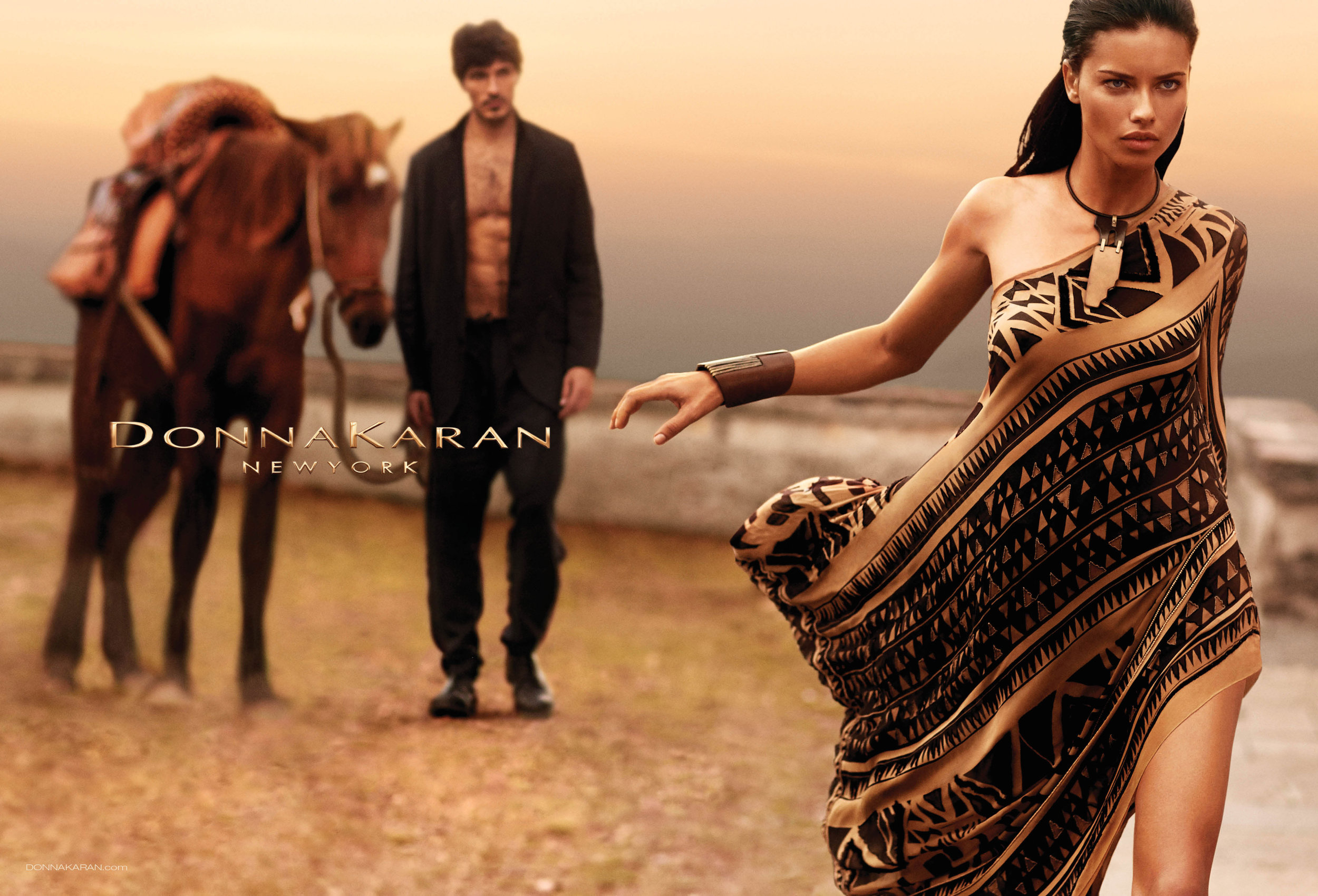 Fashion shot featuring a model in a patterned one-shoulder dress, standing outdoors with a man and horse in the background.