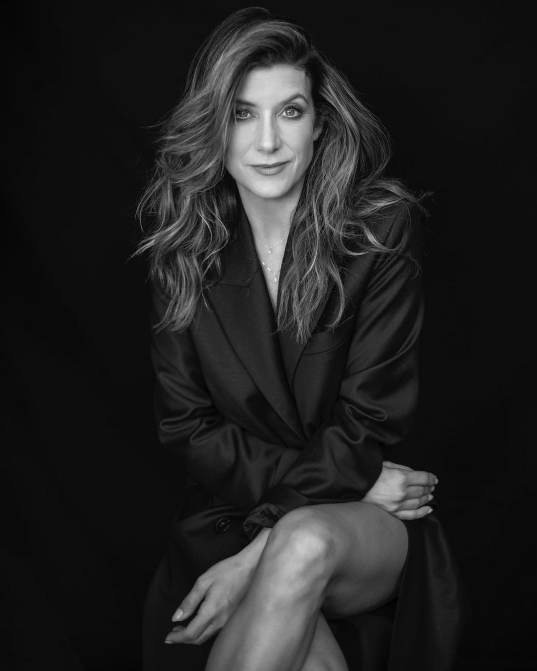 Portrait of adult woman with wavy brown hair and black silk robe seated against a dark background in a photo studio.