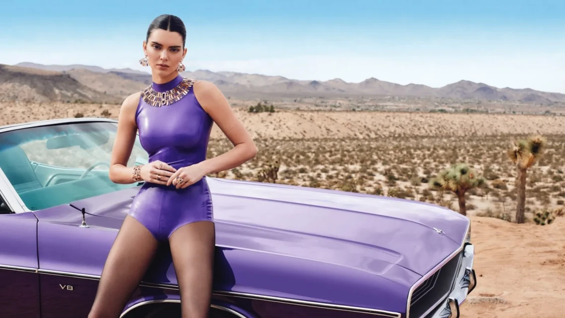 Woman in purple bodysuit poses with purple vintage car against desert backdrop, showcasing fashion, style, and automotive elements.