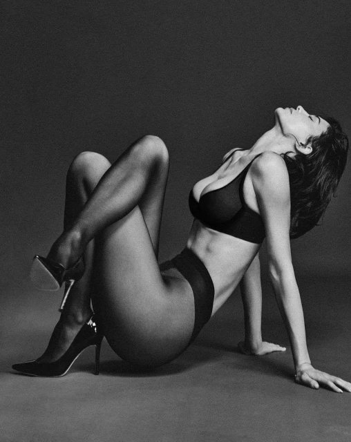 Monochrome studio shot of an adult woman in black lingerie and high heels posing with her head back and legs crossed.
