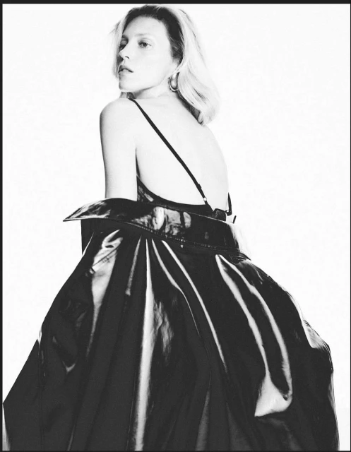 Blonde adult woman in a black dress with thin straps and a shiny black coat, looking over her shoulder in a studio shot.