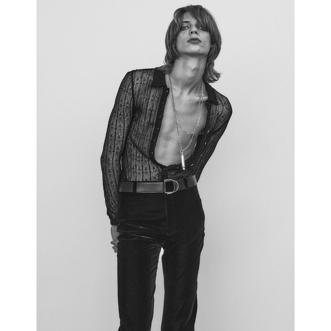Monochrome fashion portrait of a slim, fair-skinned young adult man in an unbuttoned sheer top, black pants, and silver chain necklace.