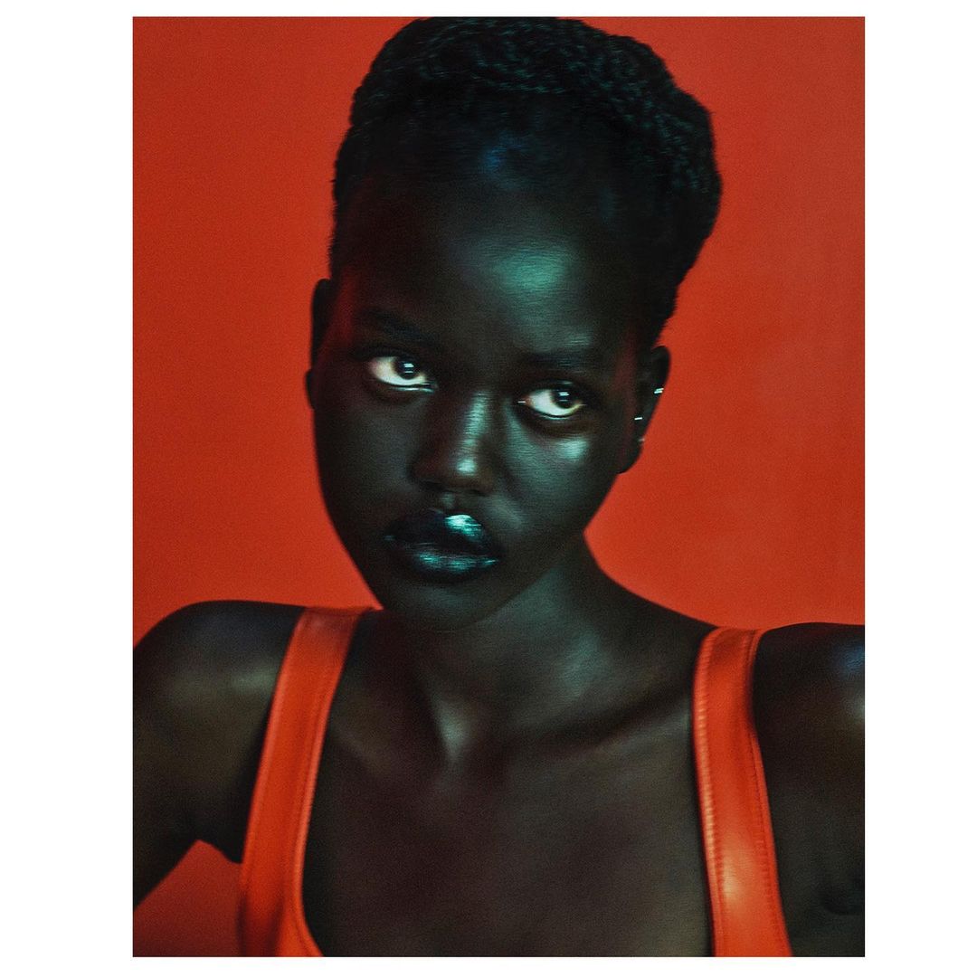 Portrait of a dark-skinned woman with glossy green lipstick, wearing a red top against a red background in a studio setting.