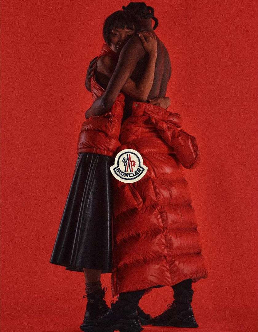 Fashion shot of a couple embracing, wearing red puffer coats and black skirt against a red backdrop, featuring the Moncler logo.