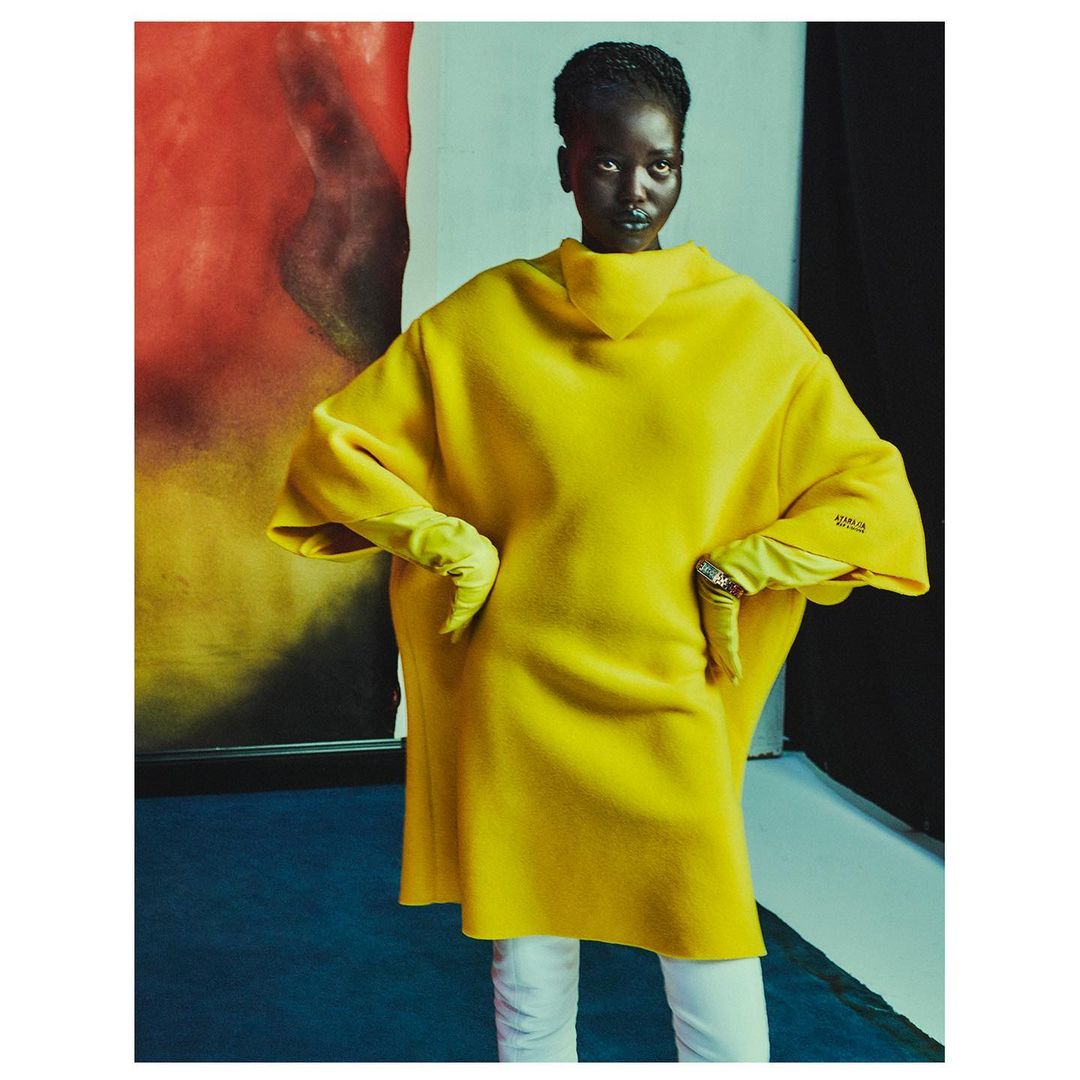 A dark-skinned woman is posing in a yellow coat, light gloves and white pants in a colorful indoor studio setting, fashion shot.