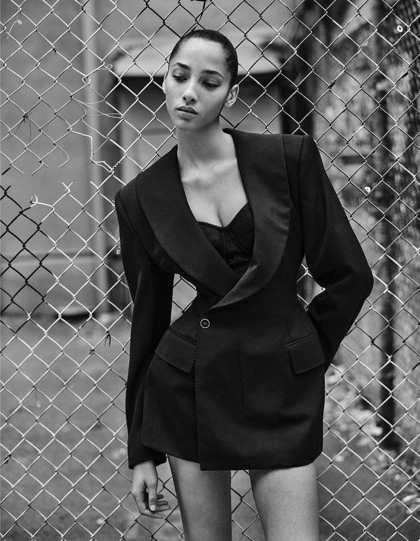 A woman in a black blazer dress stands in front of a chain-link fence in a black-and-white fashion photograph, showcasing an editorial style.