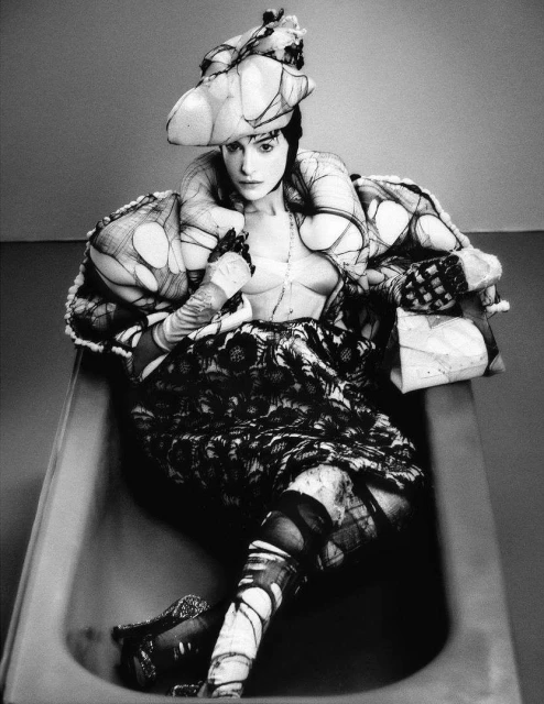 Monochrome fashion portrait of a woman in a bathtub wearing an avant-garde outfit with a sculptural hat, gloves, corset, and patterned fabrics.