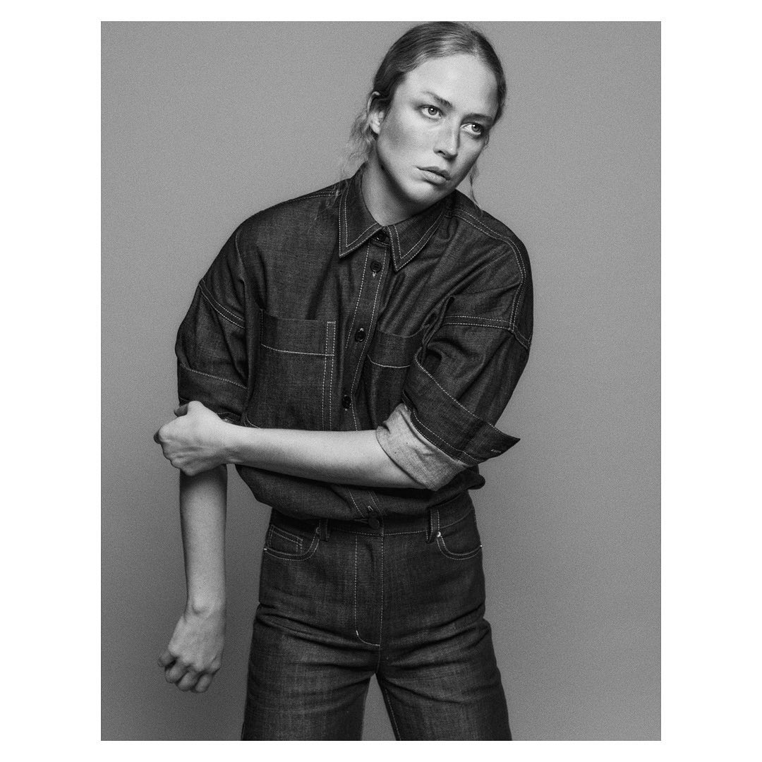Black and white portrait of a woman wearing a denim button-down shirt and trousers against a grey backdrop.