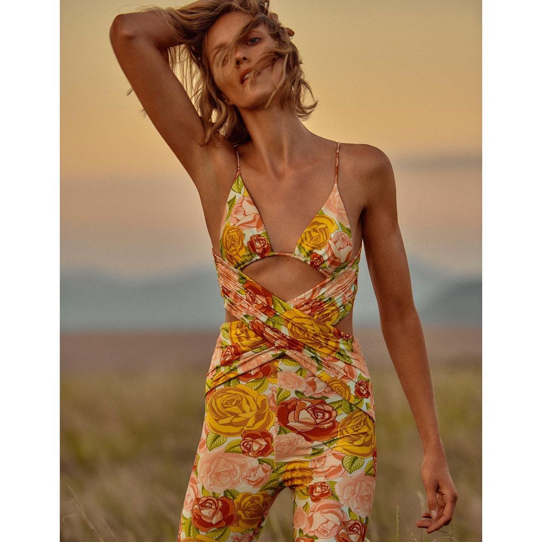 A woman in a floral print jumpsuit poses in a field against a dusky sky, her hand lifted toward her head, capturing a fashion forward moment.