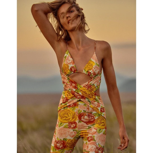 A woman in a floral print jumpsuit poses in a field against a dusky sky, her hand lifted toward her head, capturing a fashion forward moment.