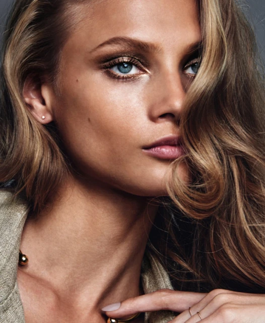 Close-up portrait of a fair-skinned adult female with wavy blonde hair, gold eye makeup, and a neutral expression in a studio setting.