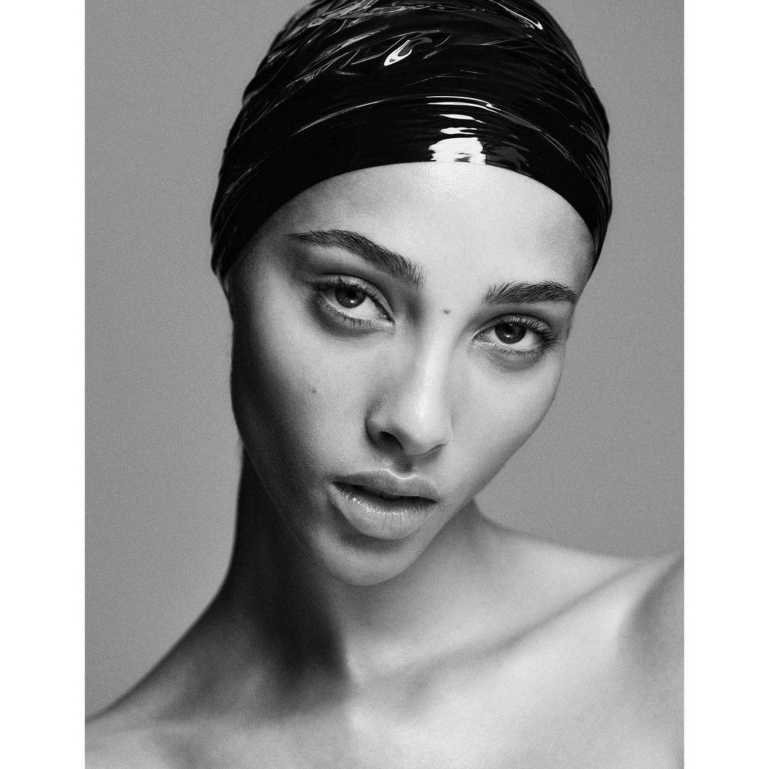 Close-up black and white portrait of a young adult woman with glossy black head covering, looking directly at the camera in a photo studio.