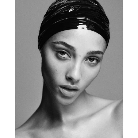 Close-up black and white portrait of a young adult woman with glossy black head covering, looking directly at the camera in a photo studio.