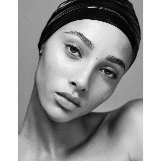 Striking black and white portrait of a woman with a sleek black head wrap, showcasing her flawless skin and captivating gaze in a minimalist studio setting.
