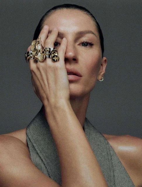 Close-up portrait of a model with her hand near her face and multiple rings, set against a gray backdrop in a beauty context.