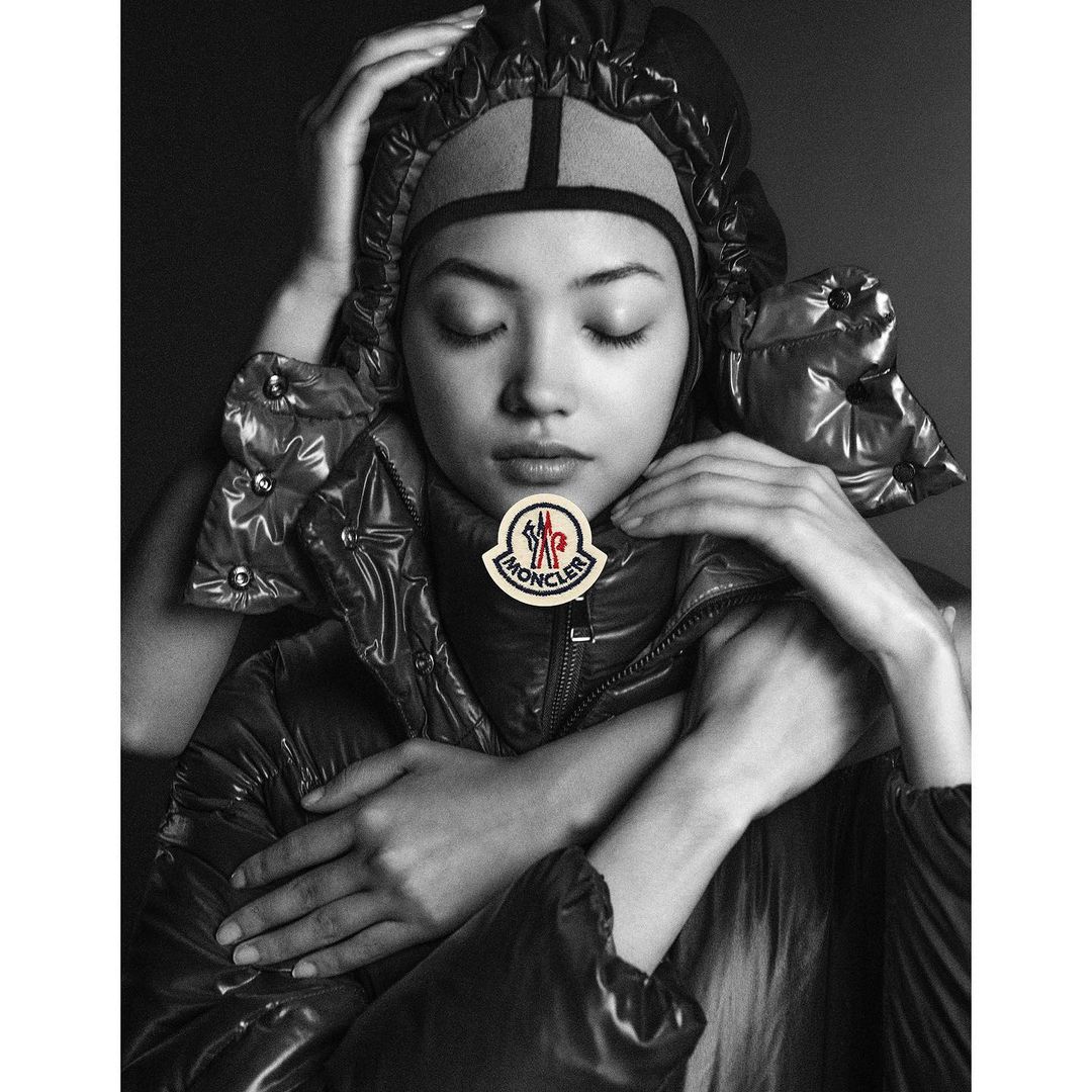 Monochrome portrait of an Asian woman with closed eyes wearing a black puffer jacket and head covering, with hands framing the face.