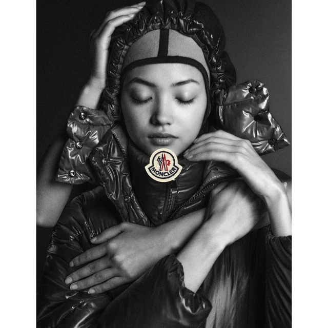 Monochrome portrait of an Asian woman with closed eyes wearing a black puffer jacket and head covering, with hands framing the face.