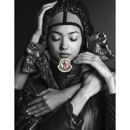 Monochrome portrait of an Asian woman with closed eyes wearing a black puffer jacket and head covering, with hands framing the face.