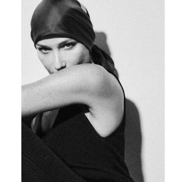 Monochrome portrait of a woman with a headscarf and dark top, posing against a light wall in a studio setting with soft shadows.