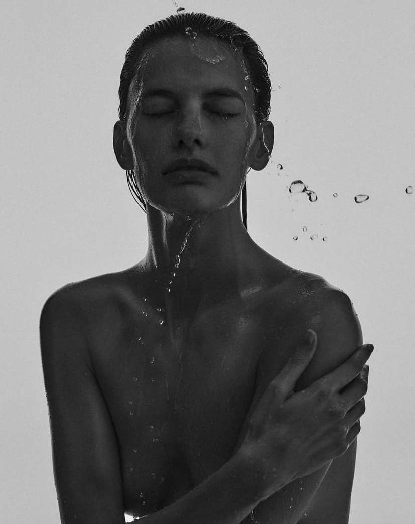 Monochrome portrait of a woman with closed eyes, water droplets on her face and shoulder, against a stark white backdrop.