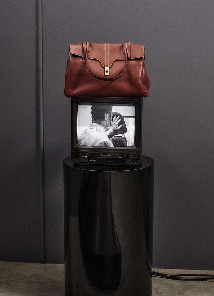 Brown leather handbag displayed on top of an older model television set on a glossy black pedestal against a grey wall.