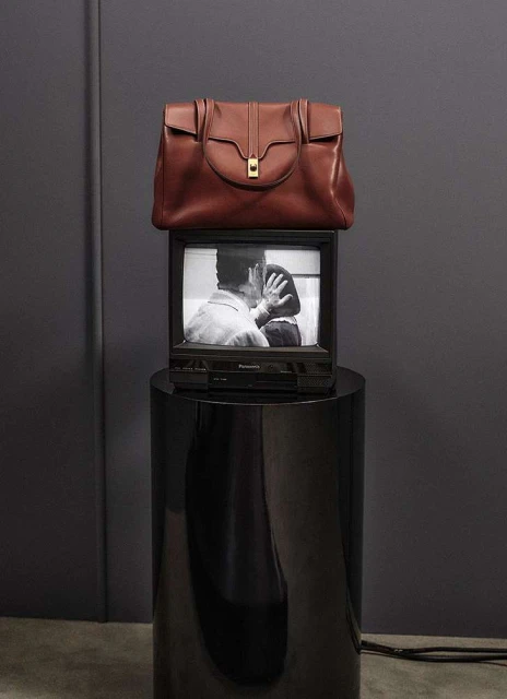 Brown leather handbag displayed on top of an older model television set on a glossy black pedestal against a grey wall.