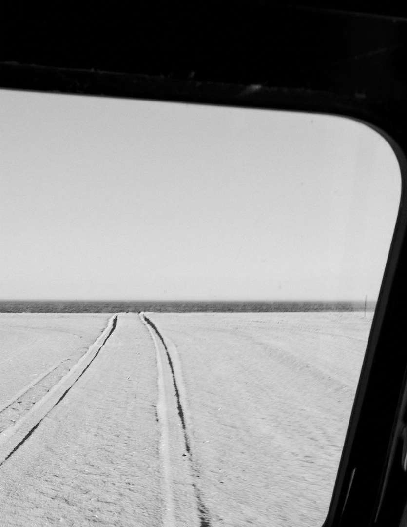 Black and white landscape shot from inside a vehicle, capturing tire tracks on a flat terrain and the horizon visible through a window.