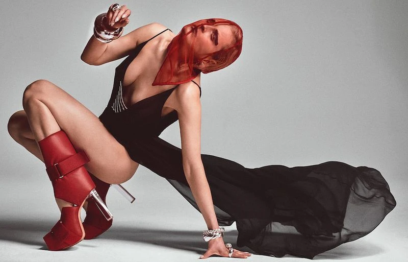 Fashion shot of a model in a black bodysuit and red boots, posing in a studio with a red transparent scarf over her head.