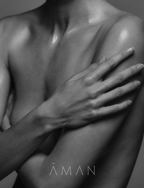 Monochrome close-up of an adult woman's bare chest and arms, skin glistens, crossed arms, logo at bottom. Studio lighting and grey background.