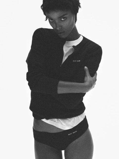 Monochrome portrait of a brunette woman wearing a black cardigan and black underwear with logo against a stark white background.