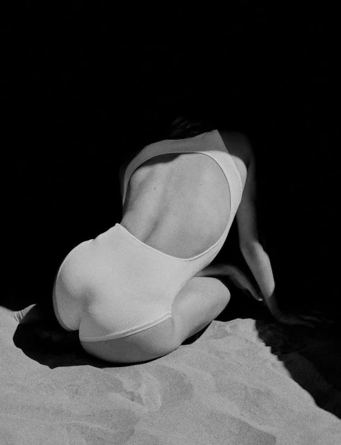Back view of adult female model kneeling on sand wearing white one-piece swimsuit against dark background in black and white photograph.