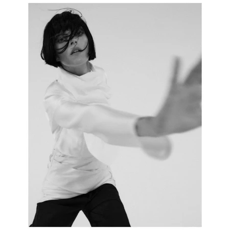 Monochrome portrait of a woman with dark bob haircut wearing a white blouse and dark trousers reaching towards camera.