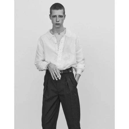 Portrait of a young man with short hair wearing a white collared shirt and dark trousers against a neutral background.