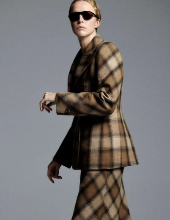 Model wearing dark sunglasses and a brown plaid blazer against a solid grey background