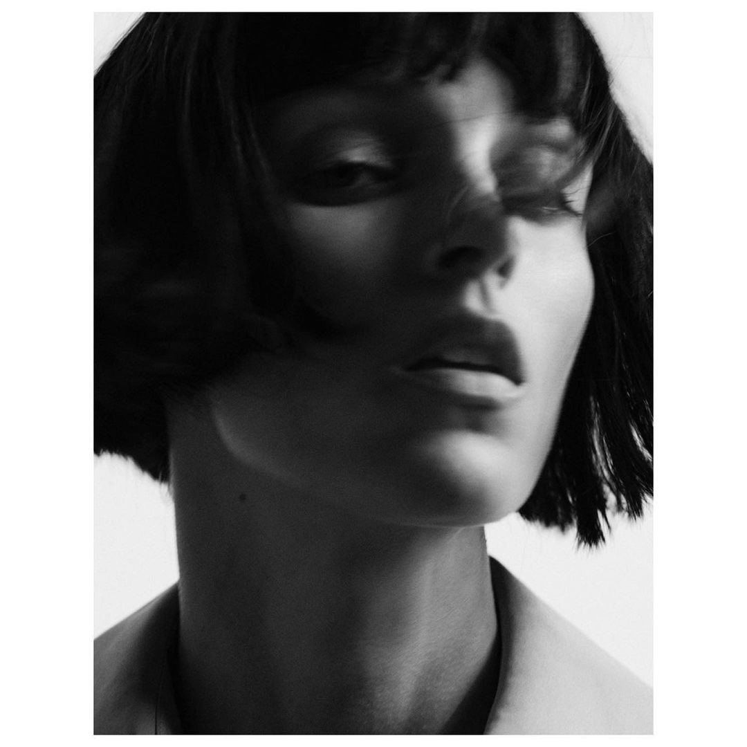 Close up monochrome portrait of a woman with dark bob hair and bangs looking downward in studio lighting.