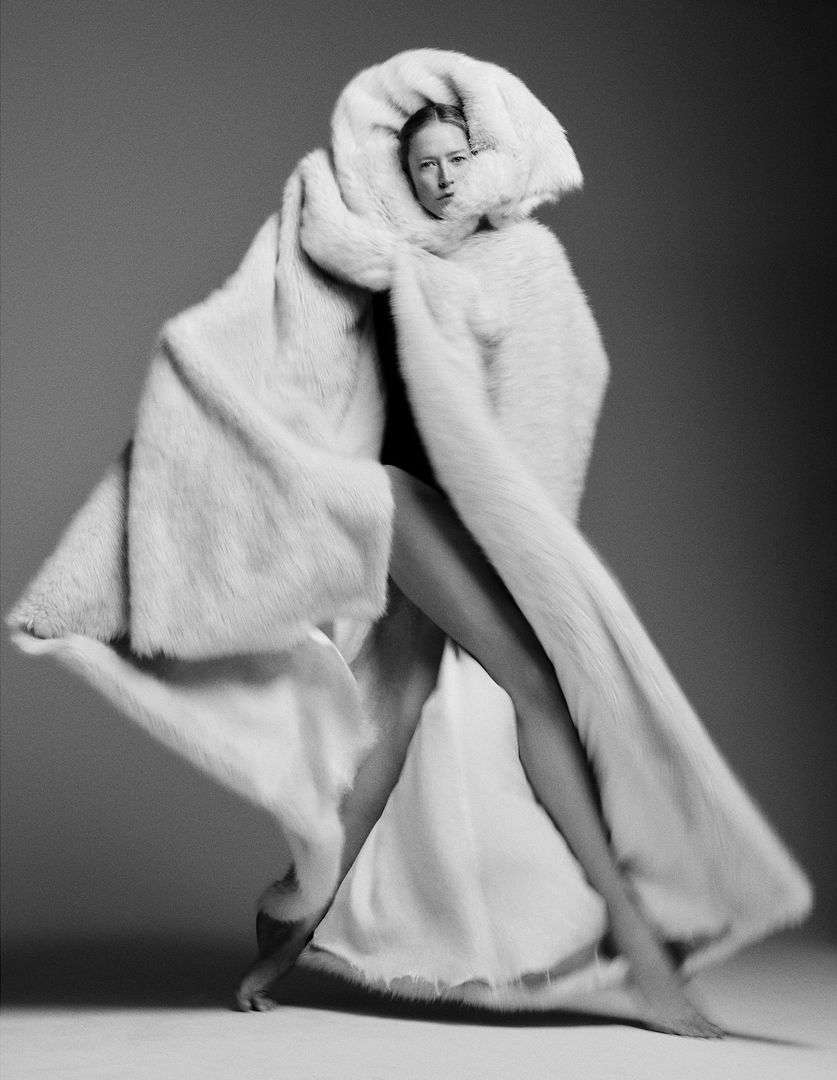 A model wearing a large white faux fur coat is striking a pose in a studio with a neutral background. The focus is on high fashion.