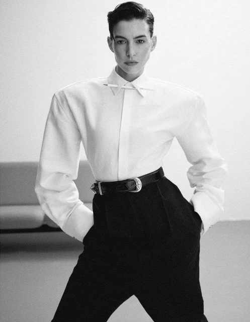 Studio shot of a woman in a white shirt and black pants with a belt, posing in front of a white background with a sofa.