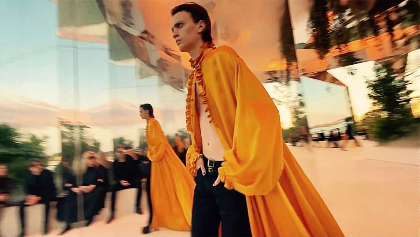 Runway show featuring male models in orange robes and black pants, audience of people, glass and sky reflections, fashion event, and outdoor setting.
