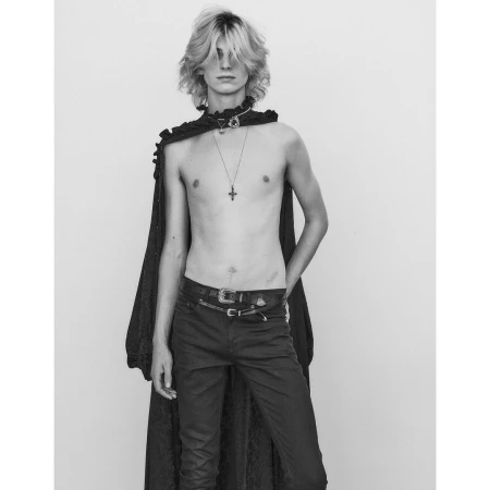 Black and white portrait of a young male model with long hair wearing a dark ruffled cape and necklaces.