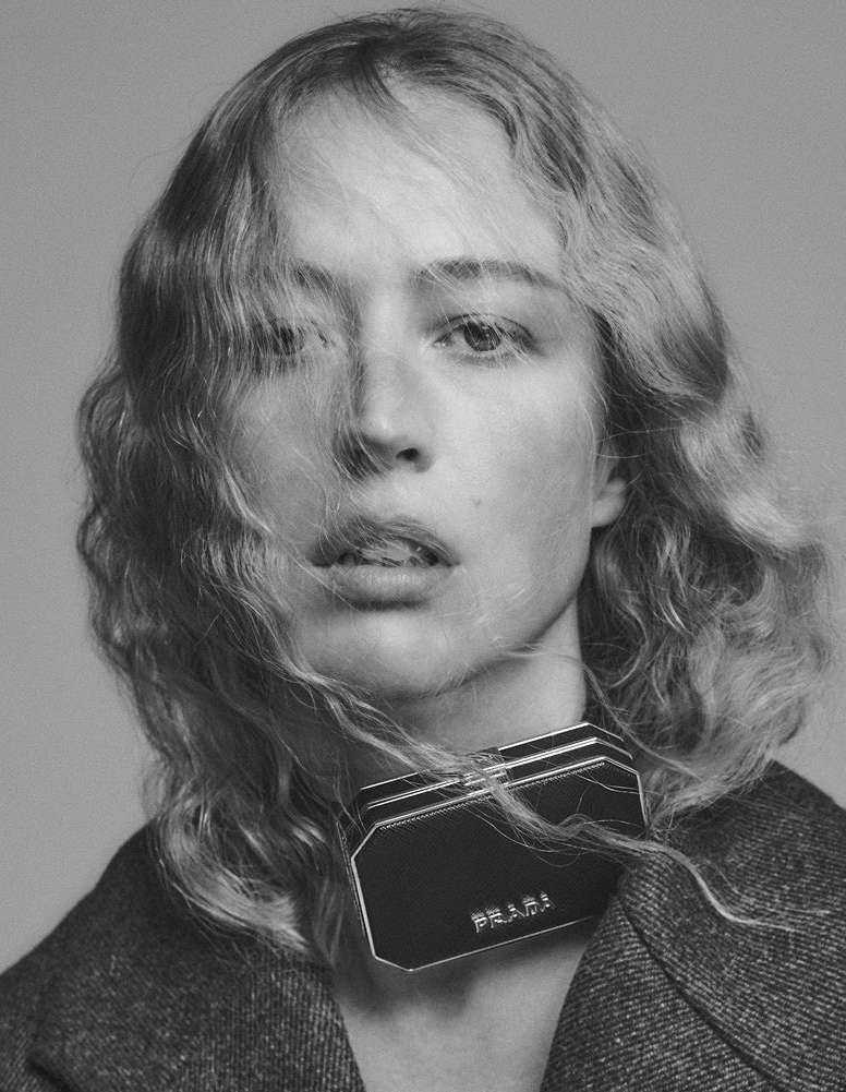 Black and white portrait of a blonde woman wearing a grey coat and black necklace featuring the PRADA logo. Close up shot.