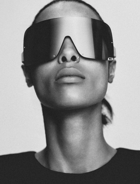 Monochrome portrait of woman wearing oversized sunglasses and a black t-shirt against a plain light background in a studio setting.