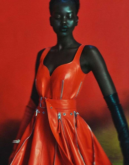 Elegant, dark-skinned woman in a striking red, belted, vinyl dress with zipper details and long black gloves against a vibrant red backdrop.